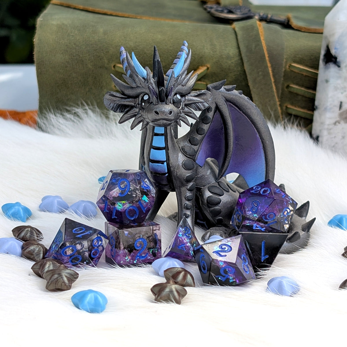 A fantasy dragon figurine in dark gray and black with bright blue accents, displayed with matching translucent blue and purple polyhedral dice on a white faux-fur surface. A large book and a white stone-like object are blurred in the background.