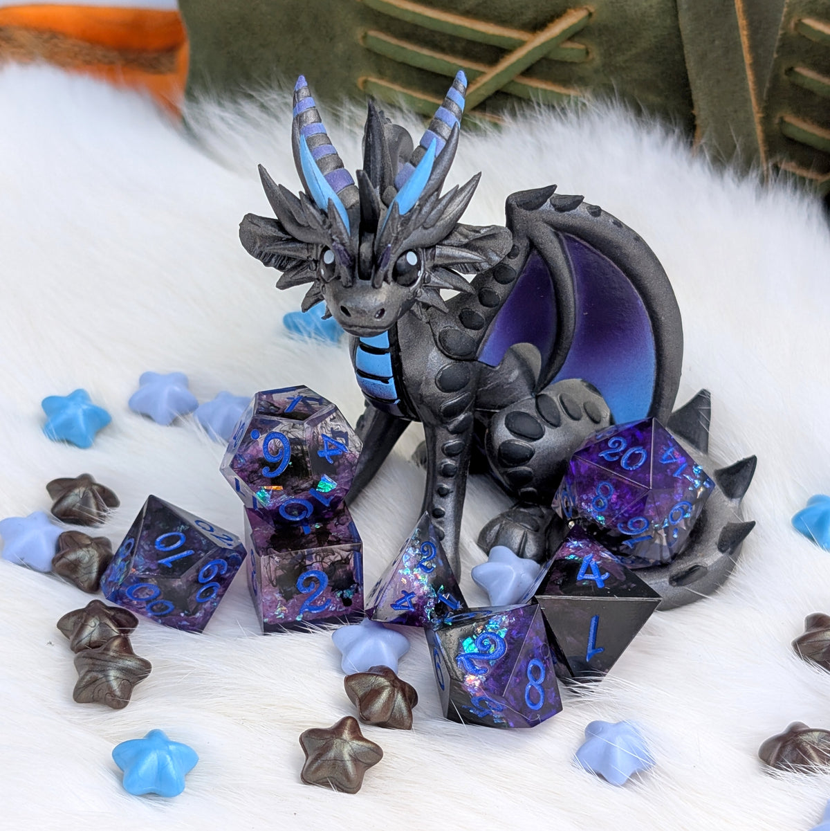 A fantasy dragon figurine in dark gray with bright blue horns and chest markings, posed among purple iridescent polyhedral dice and star-shaped dice on a white faux-fur surface.