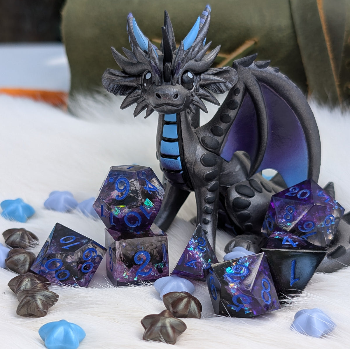A small fantasy dragon figurine in dark charcoal gray with bright blue accents, posed among matching blue and purple holographic polyhedral dice and star-shaped dice on a white faux-fur surface.