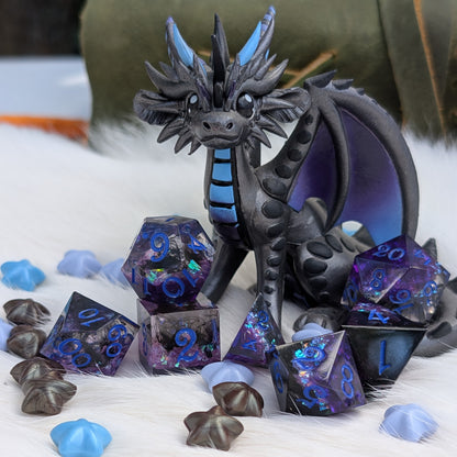 A small fantasy dragon figurine in dark charcoal gray with bright blue accents, posed among matching blue and purple holographic polyhedral dice and star-shaped dice on a white faux-fur surface.