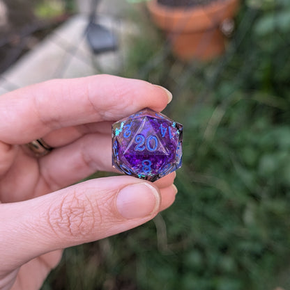 A hand holding a translucent purple and blue 20-sided d20 die outdoors with a blurred garden background.
