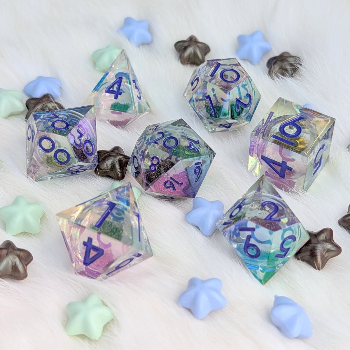 A set of translucent polyhedral gaming dice with blue numbers, decorated with colorful glitter and resin in pastel shades, arranged on a white faux fur surface with decorative star-shaped props.