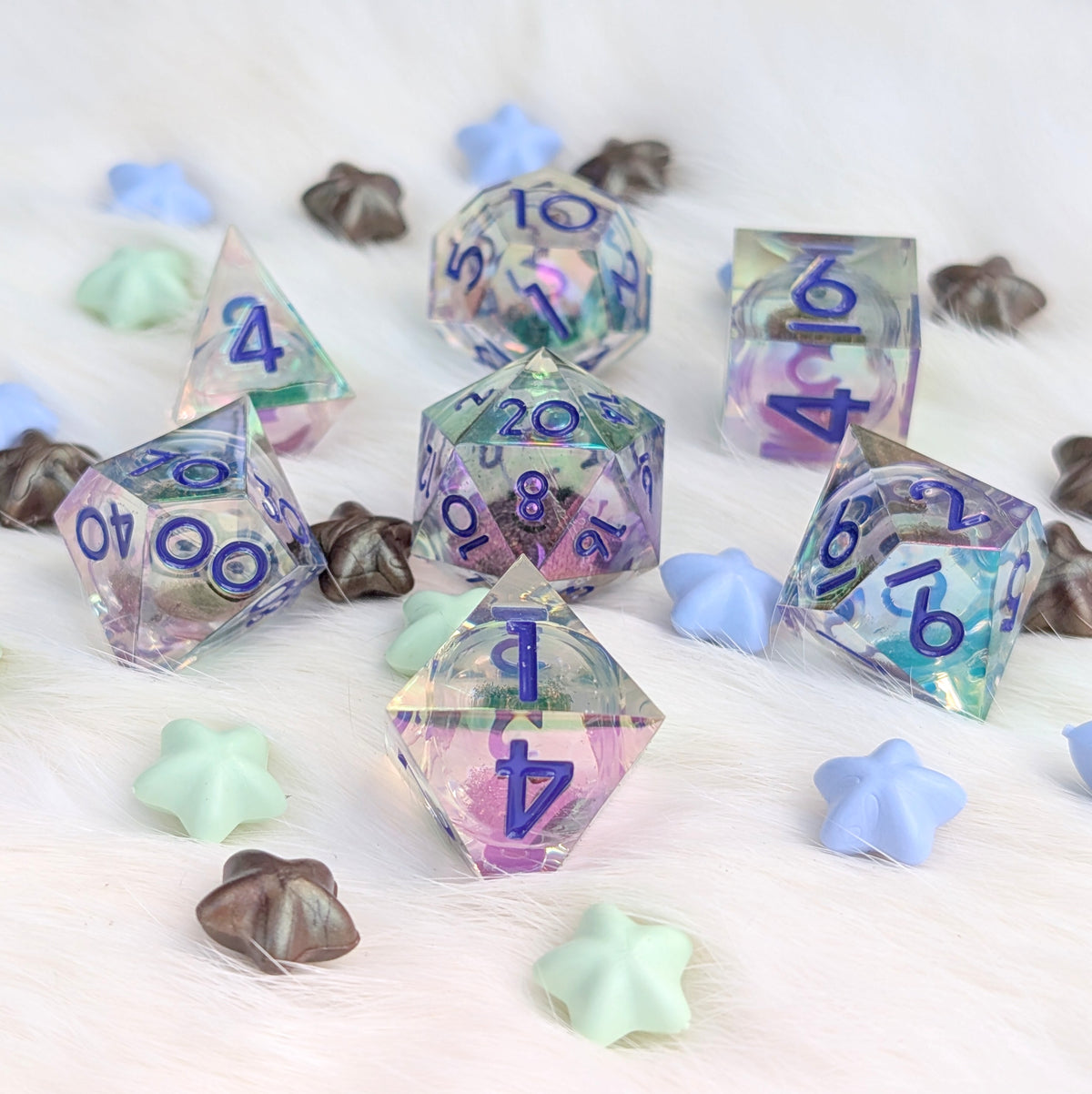 A set of translucent polyhedral gaming dice with iridescent pastel colors and blue numbers, arranged on a white faux-fur surface with small decorative star and flower props.