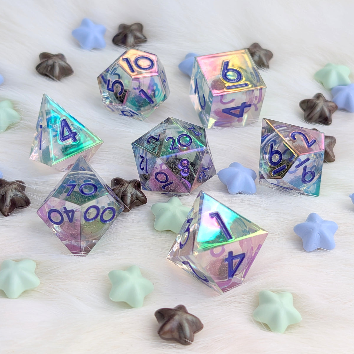 A set of holographic polyhedral gaming dice with purple numbering, scattered on a white faux fur surface with pastel star-shaped candies.