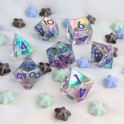 A set of holographic polyhedral gaming dice with purple numbering, scattered on a white faux fur surface with pastel star-shaped candies.