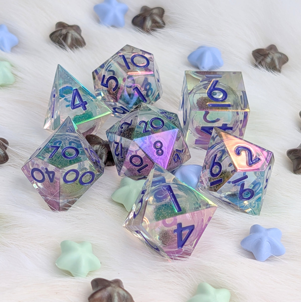 A set of holographic polyhedral gaming dice with blue printed numbers, arranged on a white faux fur surface with pastel star-shaped and shell-shaped decorative props.