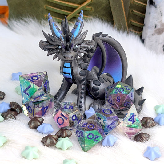 A fantasy dragon figurine in dark gray with bright blue horns and chest markings, posed among translucent polyhedral gaming dice with iridescent colors and blue numbers, on a white faux-fur surface with small decorative star-like pieces.