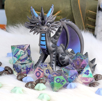 A fantasy dragon figurine in dark gray with blue accents, posed among translucent iridescent polyhedral dice on a white faux-fur surface with small pastel stones and a blurred olive-green background.