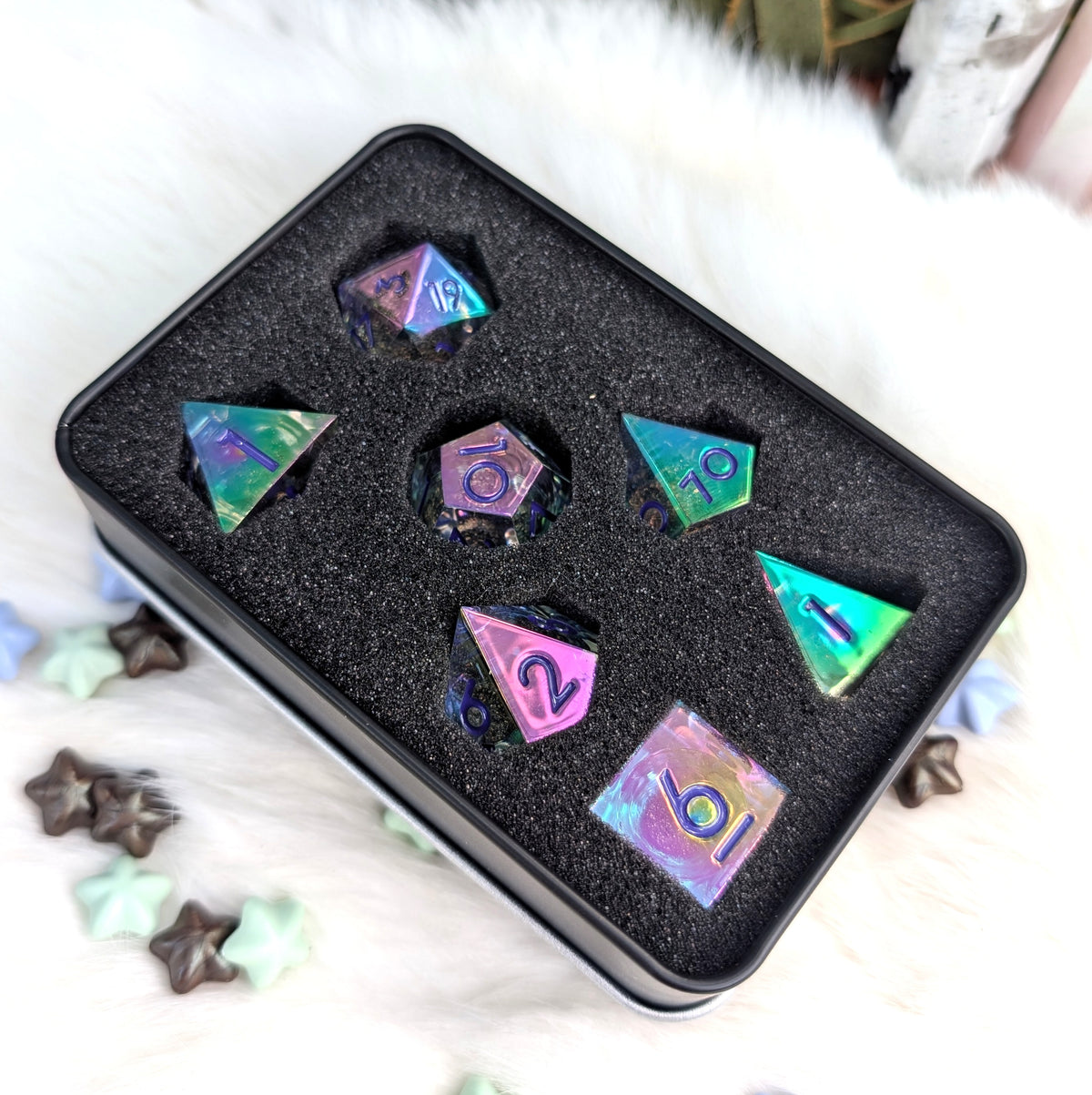 A set of holographic polyhedral dice displayed in a black foam tray inside a square metal tin, placed on a white faux-fur surface with small star-shaped props.