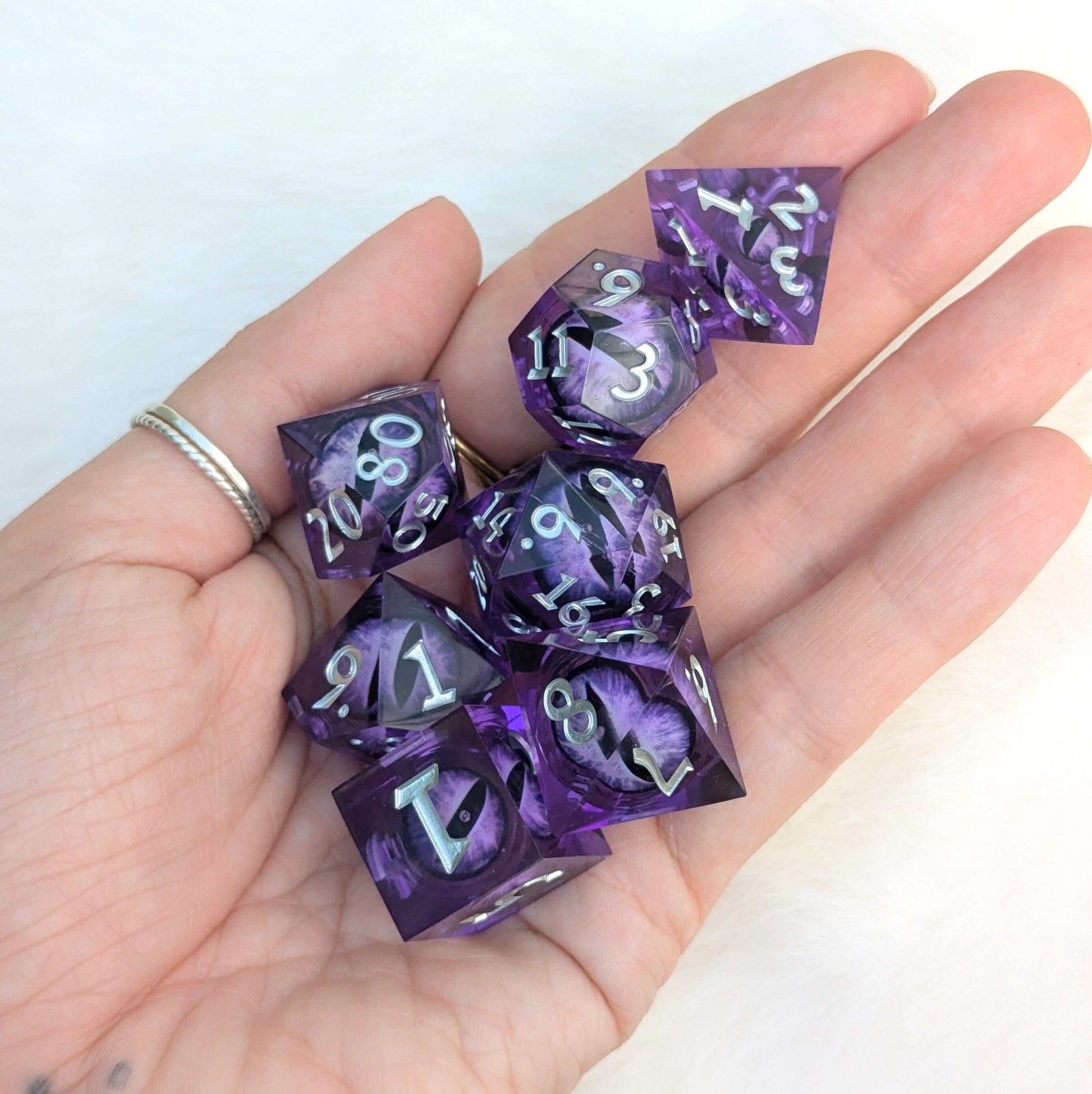 Hand holding 7-piece All-Seeing Eye Dice Set in Amethyst Edition with purple liquid core and sharp edge resin design.