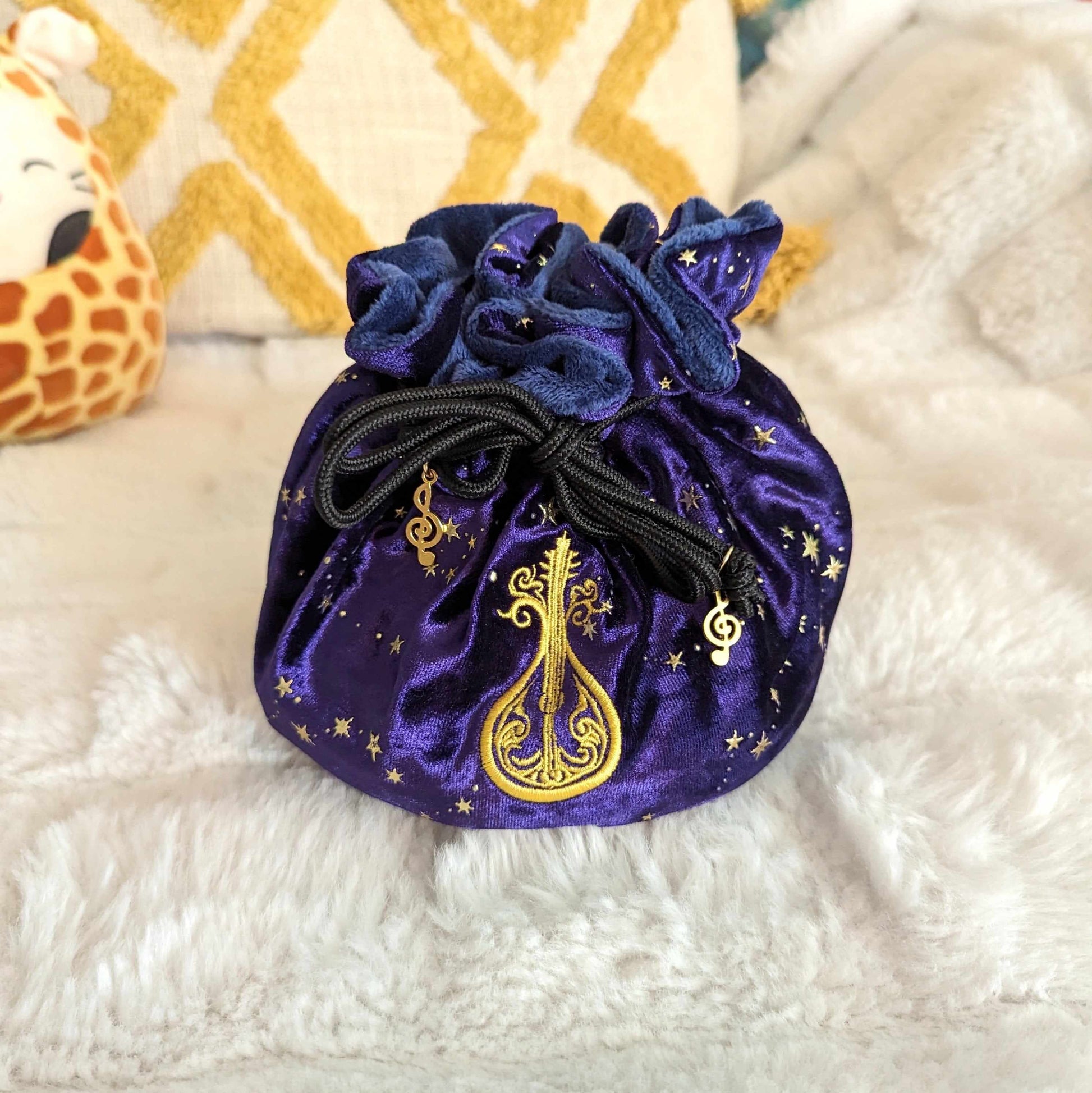 Purple velvet bard dice bag with embroidered lute design and musical charm drawstrings on a plush surface.