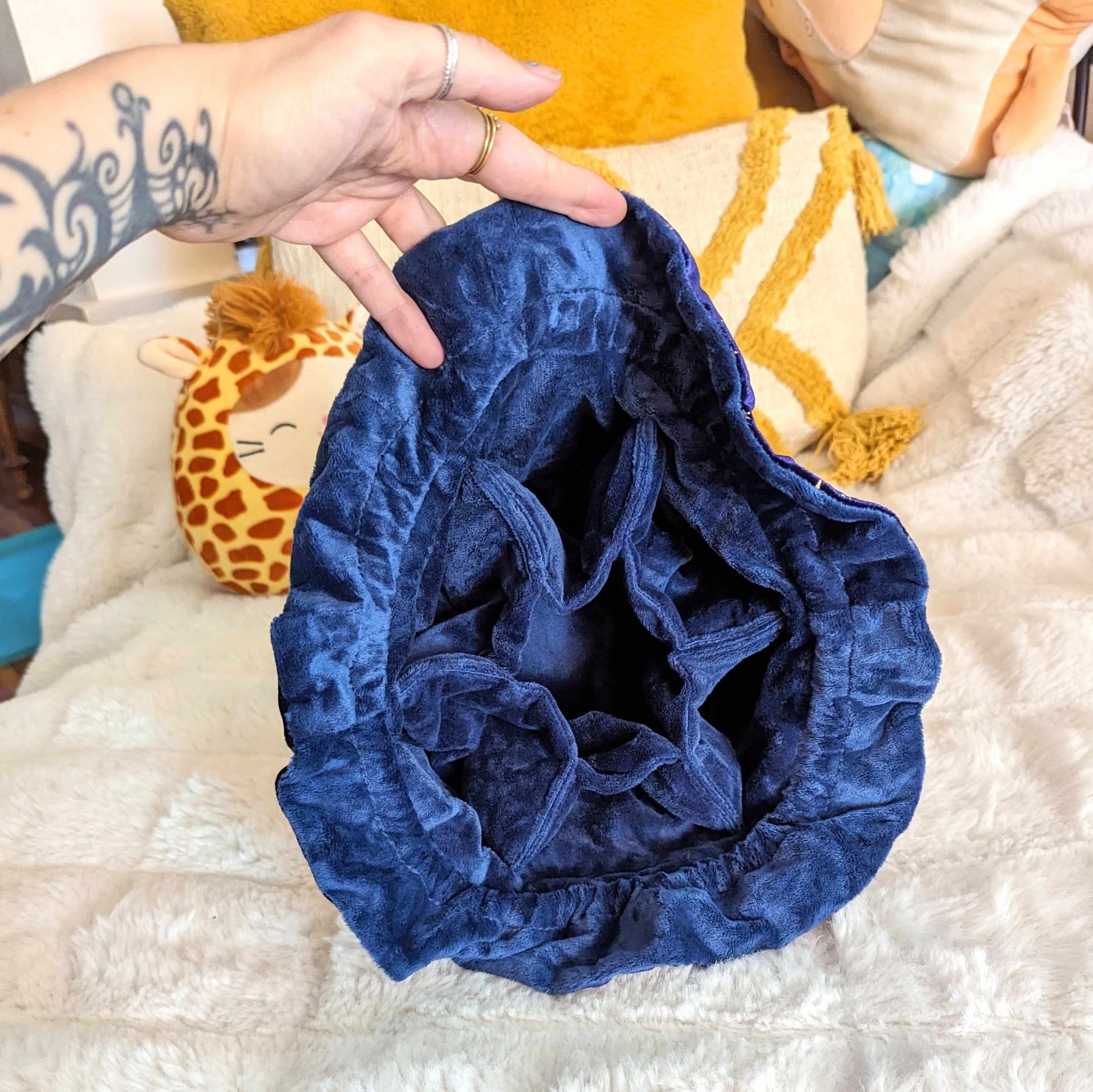 Open multi-pocket Bard dice bag in blue velvet fabric held by hand, showing interior space on a cozy bed setting.