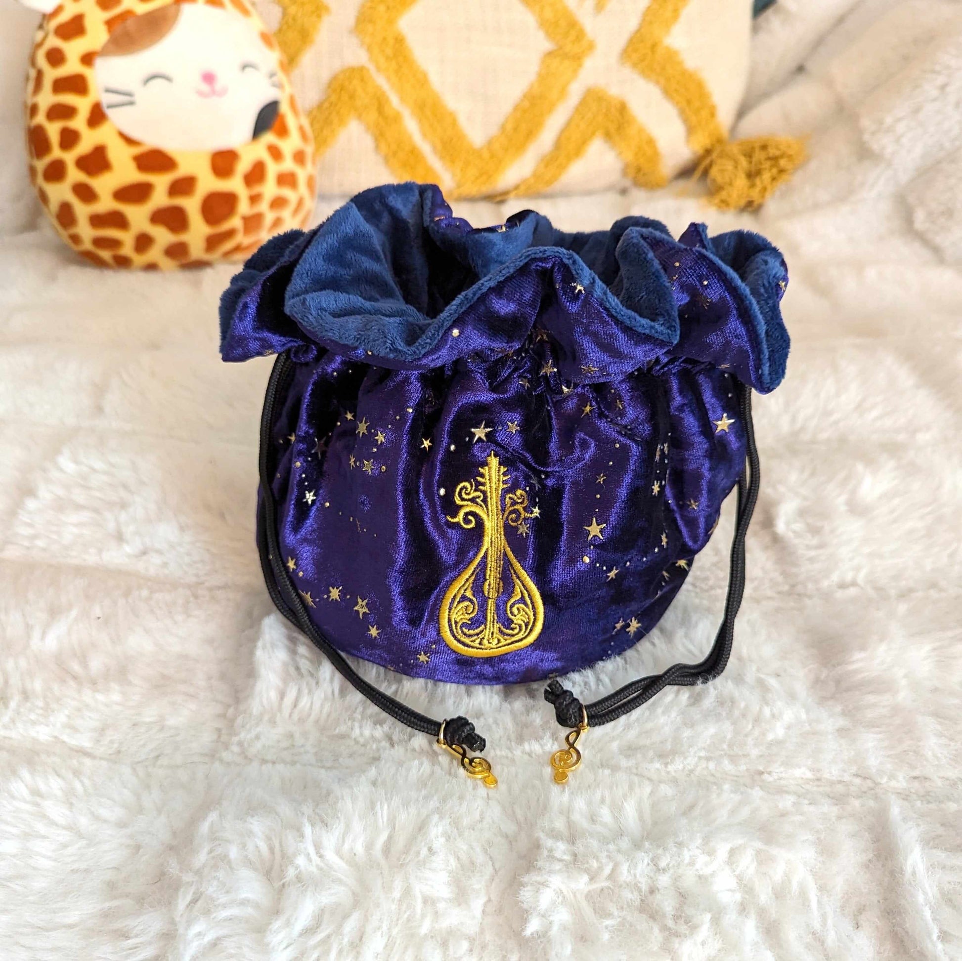 Purple velvet bard dice bag with embroidered lute, featuring musical charm drawstrings, on a cozy fabric background.