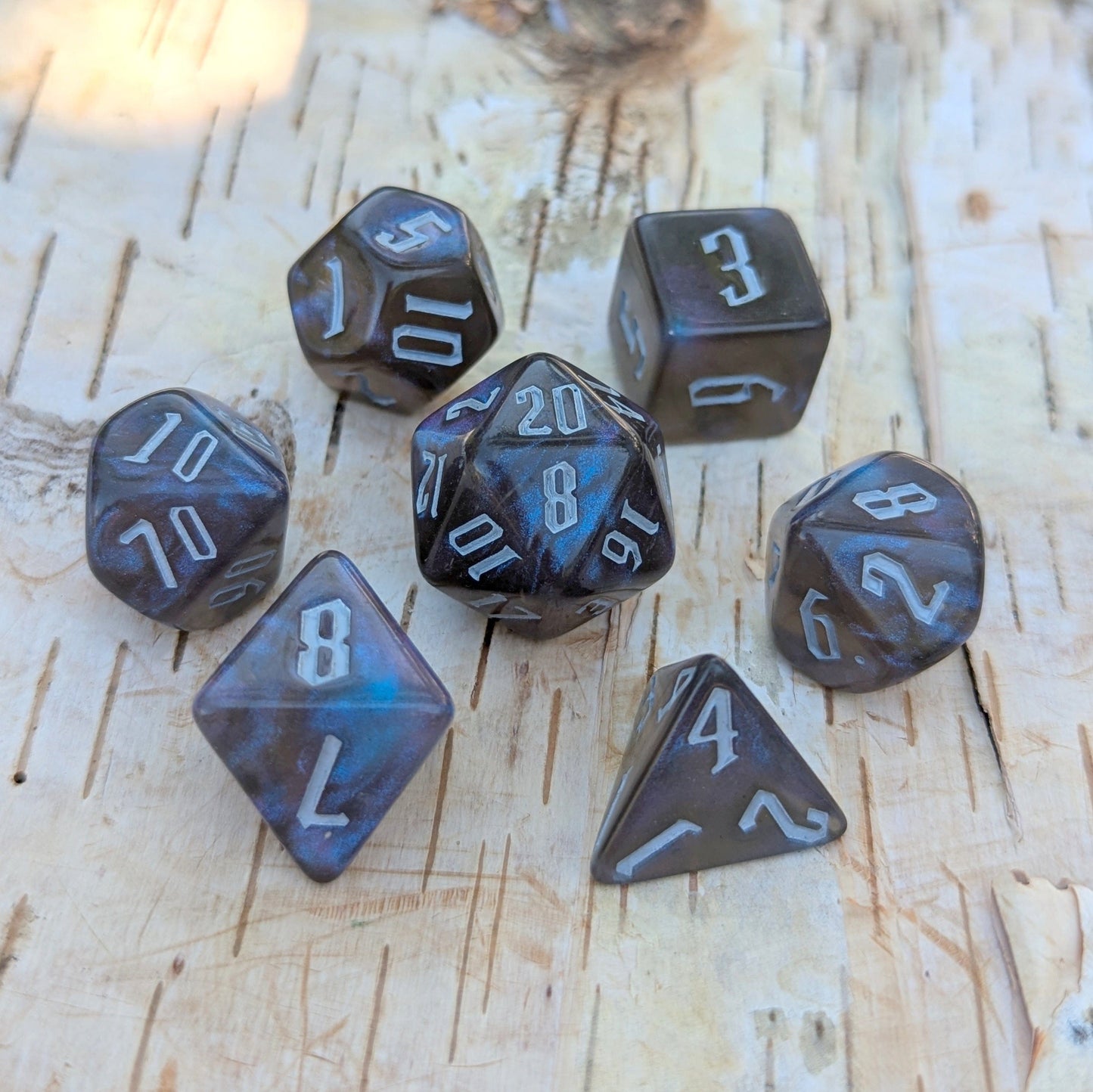 Black and blue glitter acrylic dice set with fantasy font on a wooden surface, 7-piece polyhedral set for tabletop RPGs like DnD.