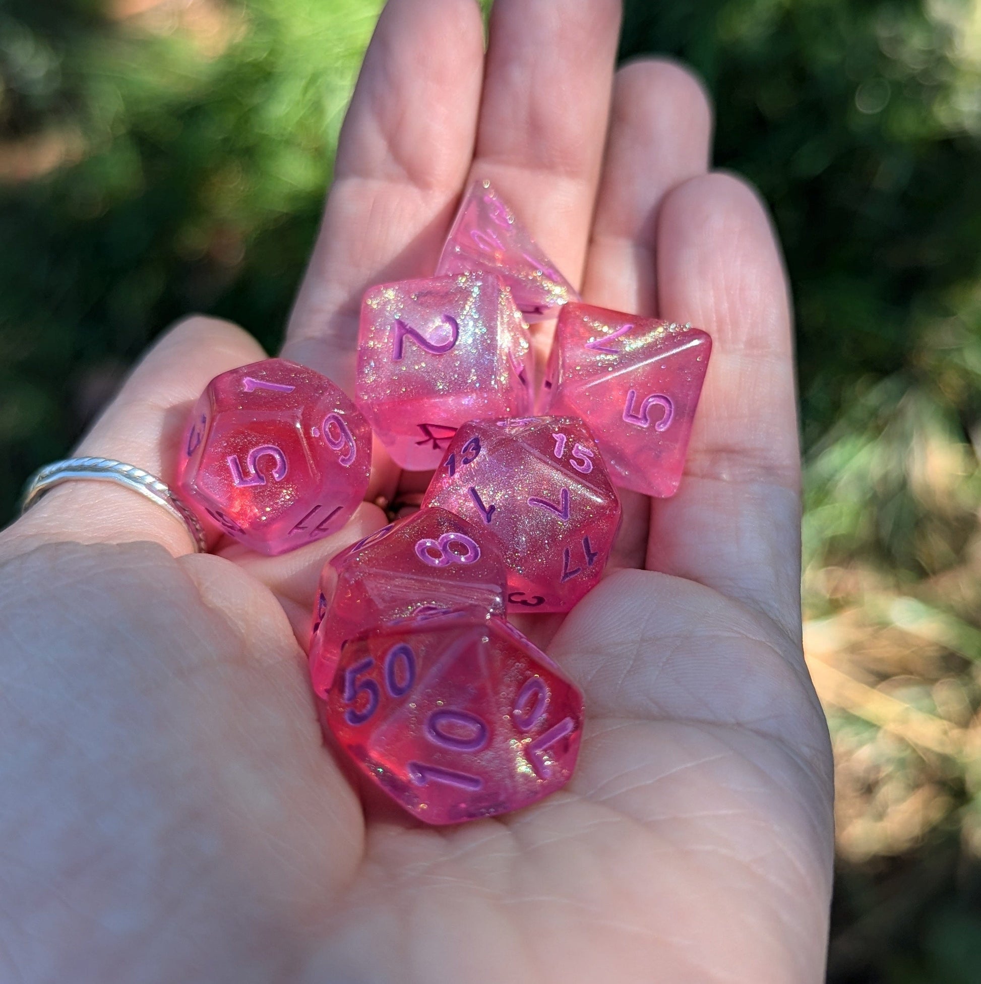 Hand holding pink glitter Cheshire Will O Wisp Dice Set for TTRPG, featuring 7 shimmering polyhedral dice with purple numbers.