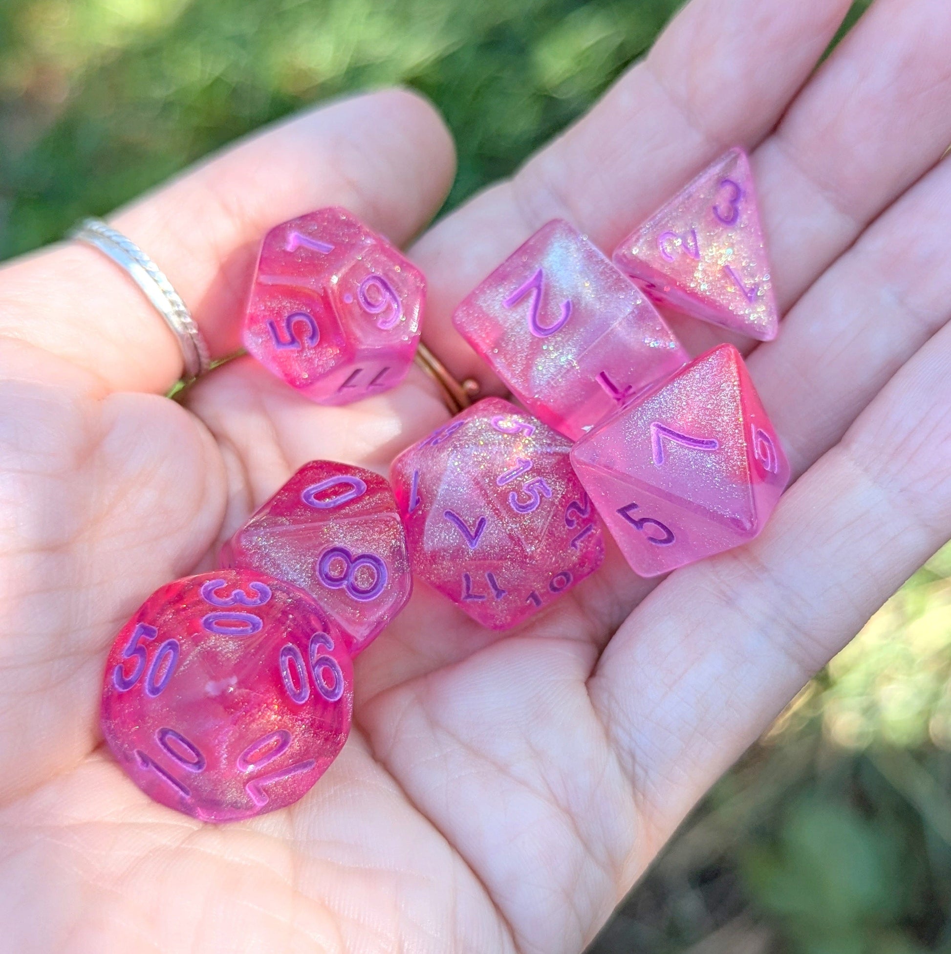 Hand holding Cheshire Will O Wisp Dice Set, bright pink with purple numbers, shimmering acrylic polyhedral TTRPG dice for DnD.