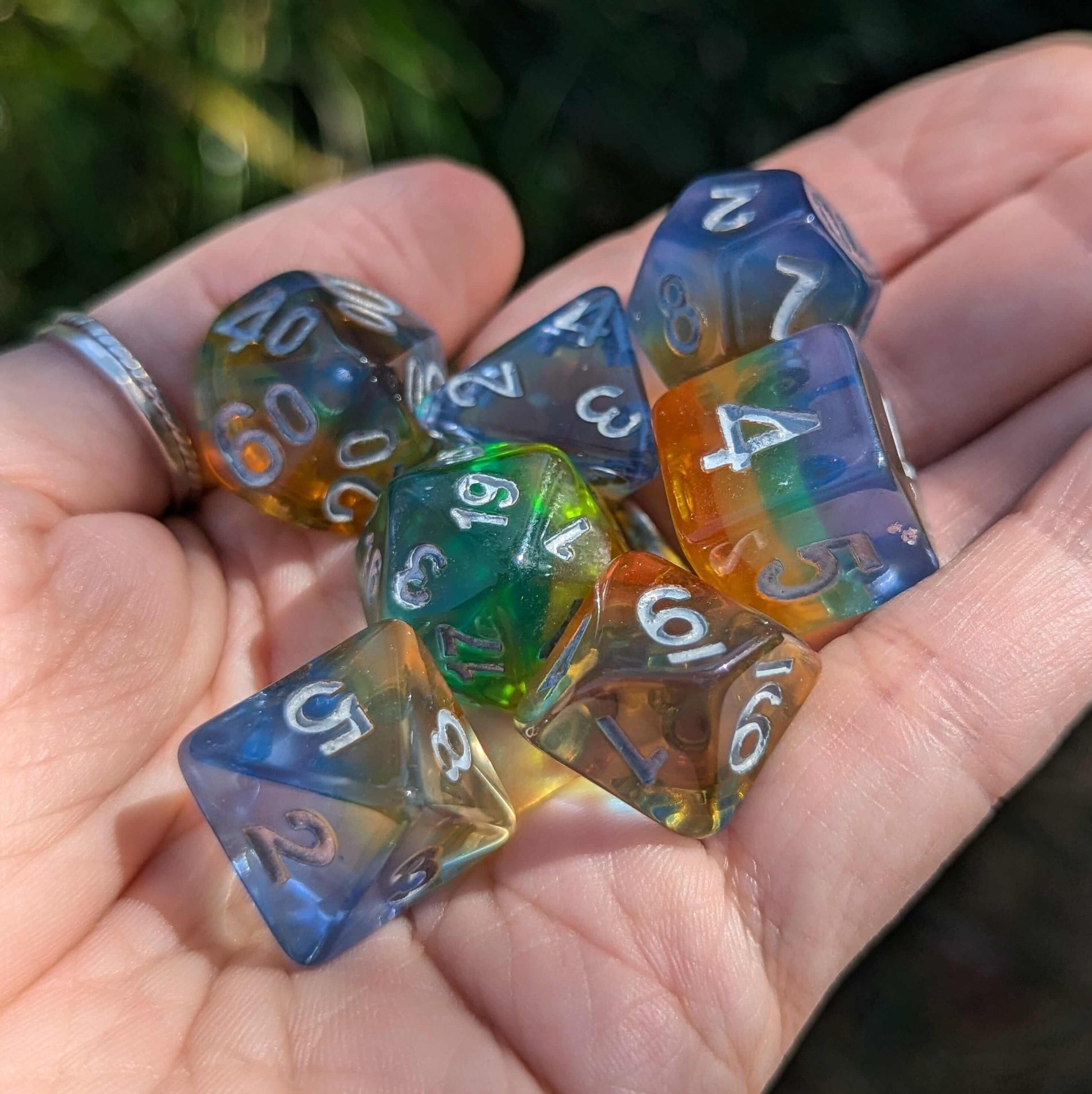 Hand holding rainbow-layered TTRPG dice set with blue, green, yellow, and orange colors, silver numbers visible in sunlight.