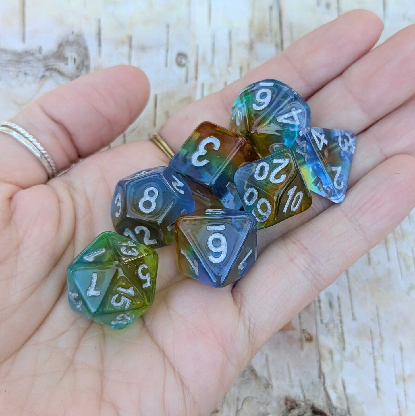 Hand holding rainbow cocktail dice set with silver font for tabletop RPGs, featuring vibrant blue, green, yellow, and orange colors.