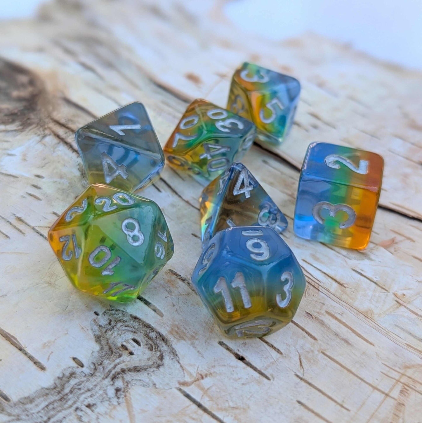 Rainbow layered TTRPG dice set with silver numbers, displaying vibrant blue, green, yellow, and orange colors on a wooden surface.