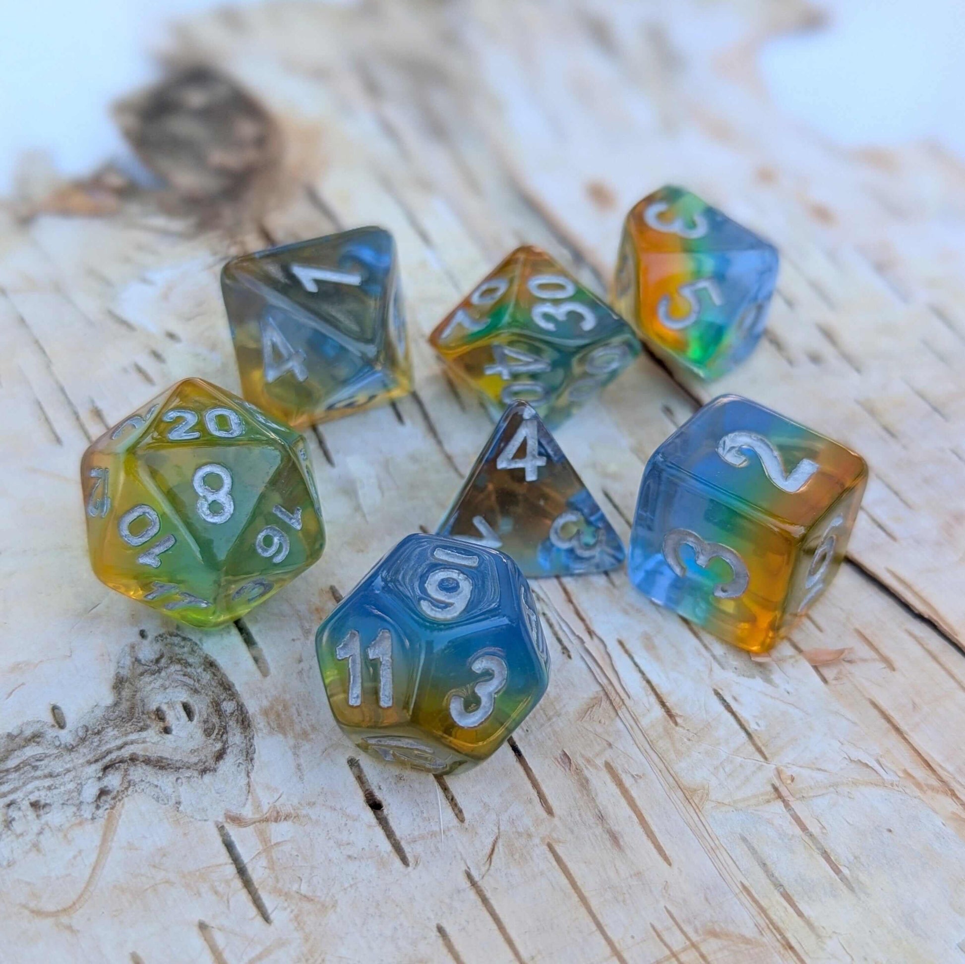 Rainbow layered TTRPG dice set with blue, green, yellow, orange shades and silver numbers on a wooden surface.