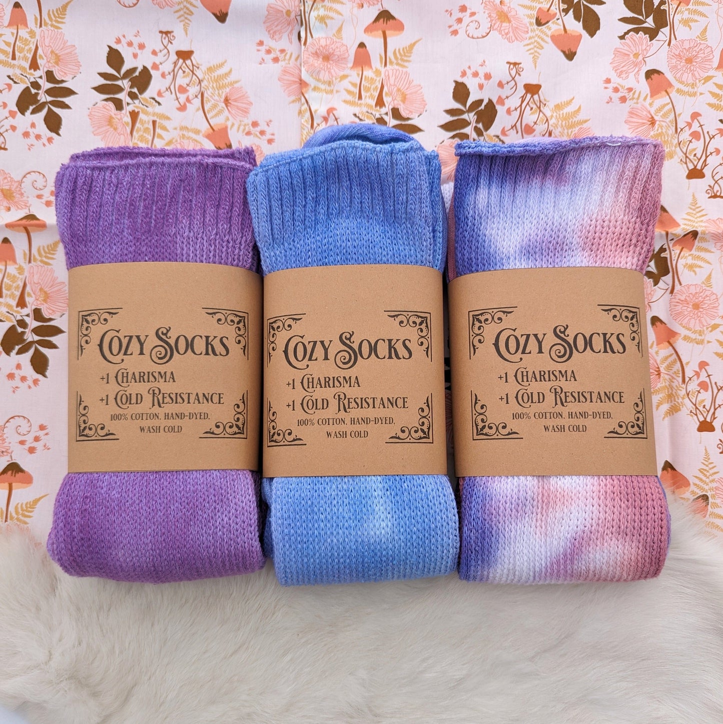 Three pairs of Cozy Sea Elf Socks in purple, blue, and tie-dye, made from 100% hand-dyed cotton, on a floral background.