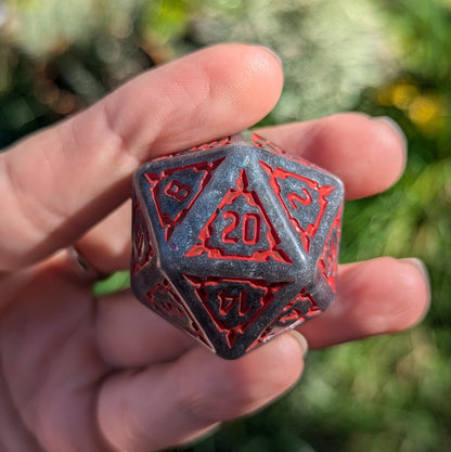 Hand holding a dark grey glittering 20-sided dice with red numbers from the extra large Dark Castle Dice set.