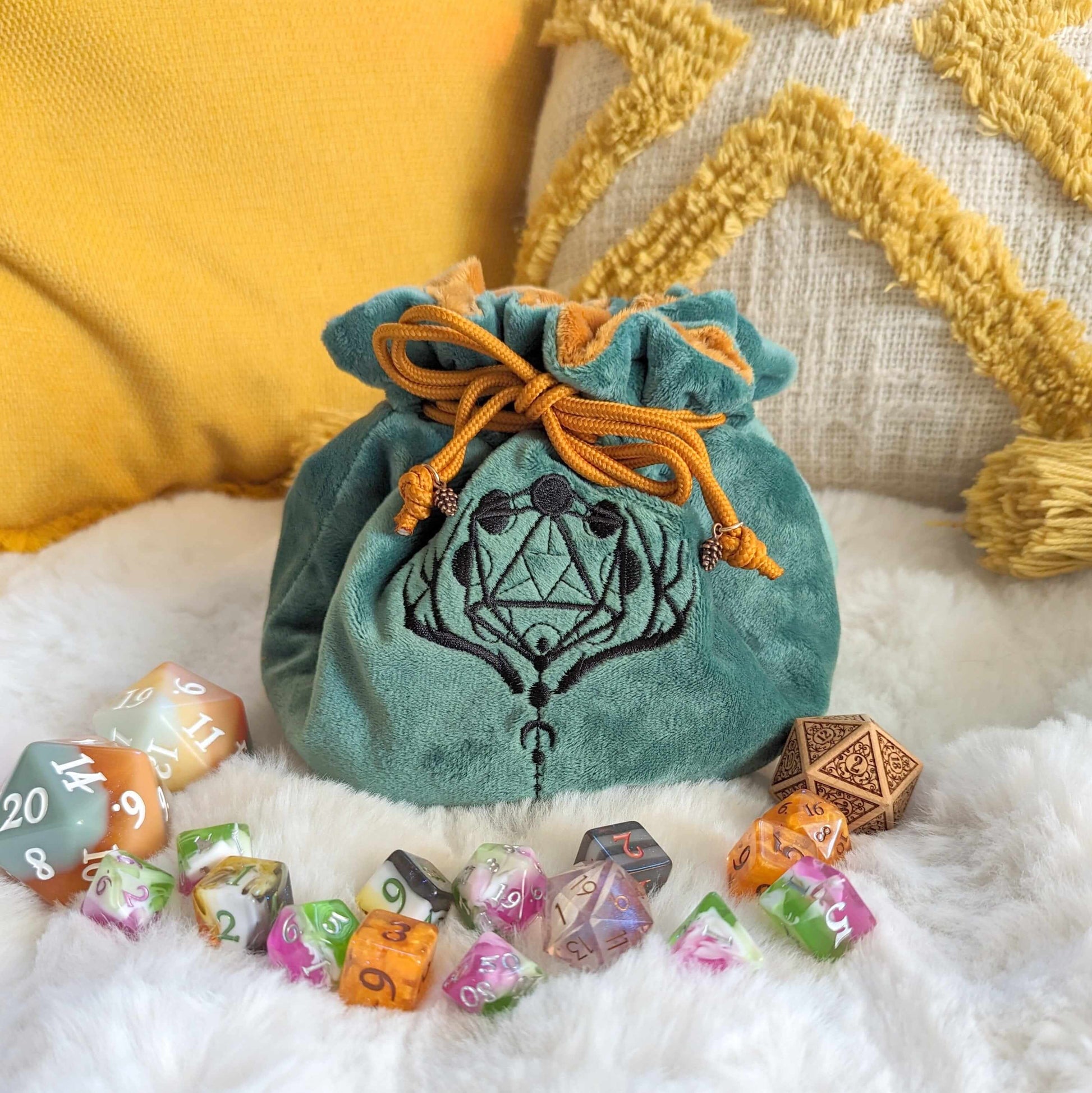 Green and brown druid dice bag with embroidered D20 and moon design, featuring drawstrings with pinecone beads, surrounded by colorful dice.