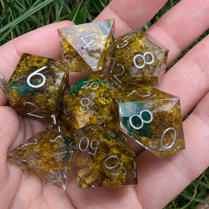 Hand holding the Enchanted Meadow Sharp Edge Dice Set with yellow flowers and green sprigs in clear resin, featuring silver numbers.