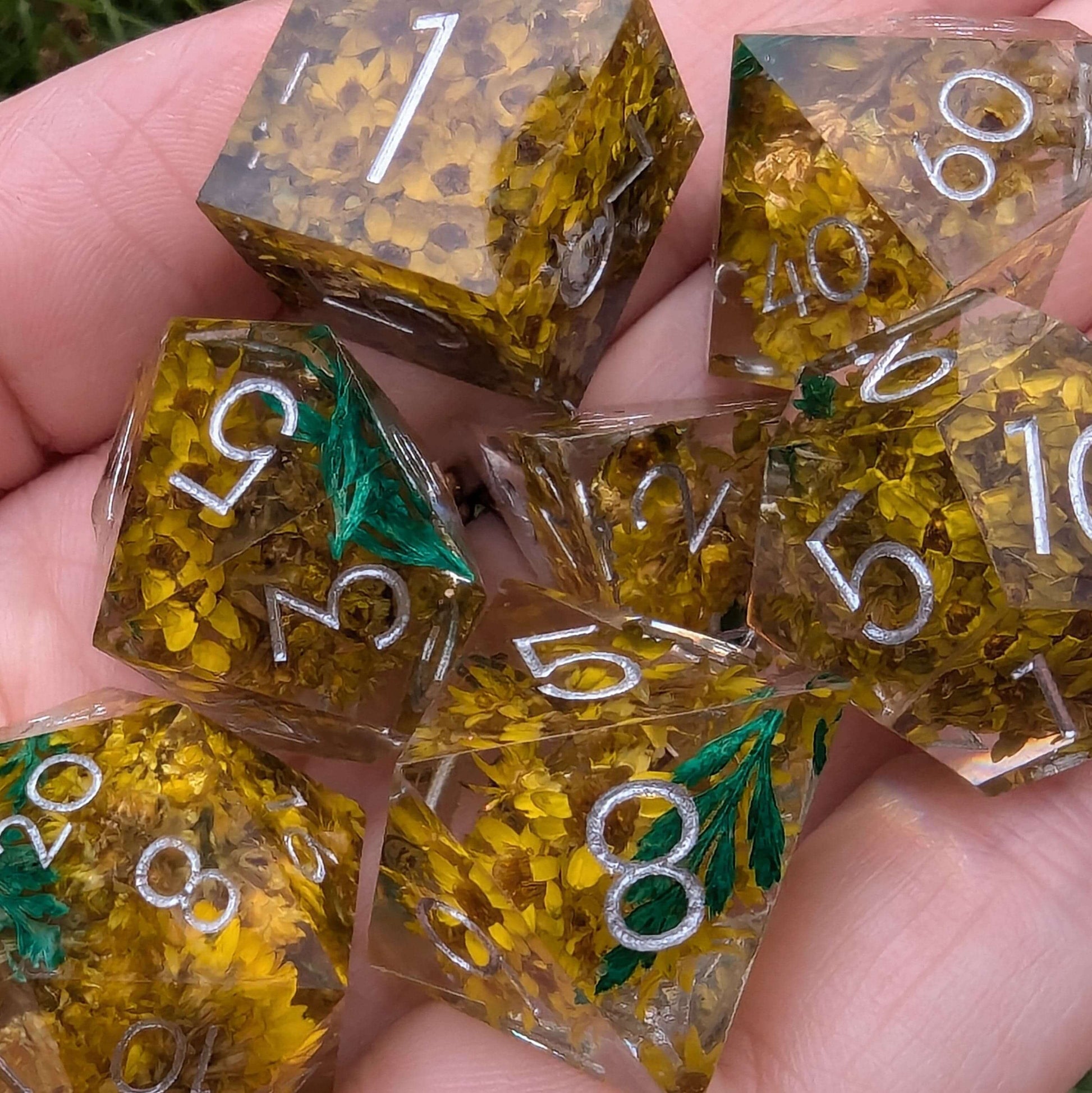 Enchanted Meadow sharp edge DnD dice set with yellow flowers and green sprigs in resin, silver numbers, handheld view.