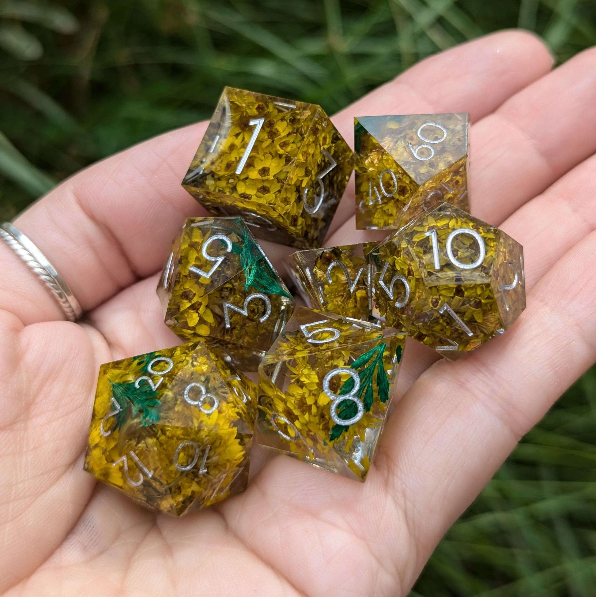 Hand holding Enchanted Meadow DnD dice set, featuring yellow flowers and green sprigs in clear resin with silver numbers.