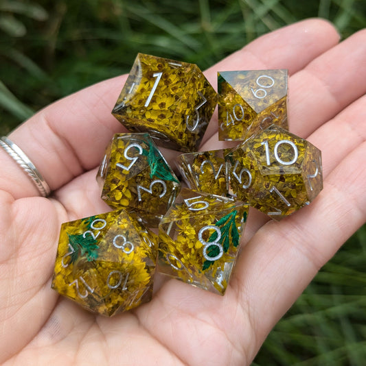 Hand holding Enchanted Meadow DnD dice set, featuring yellow flowers and green sprigs in clear resin with silver numbers.