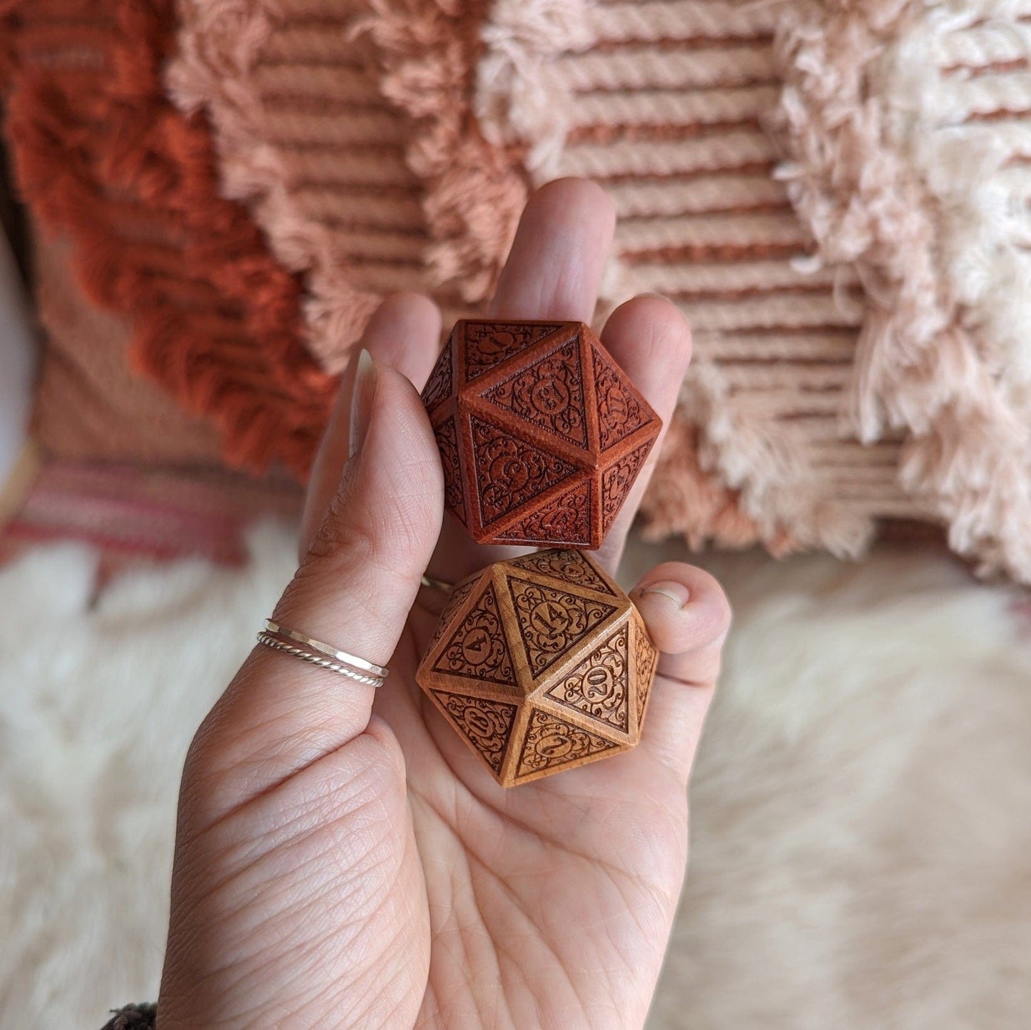 Hand holding two wood D20 dice with intricate filigree designs, one in deep red sandalwood and the other in light beech wood.