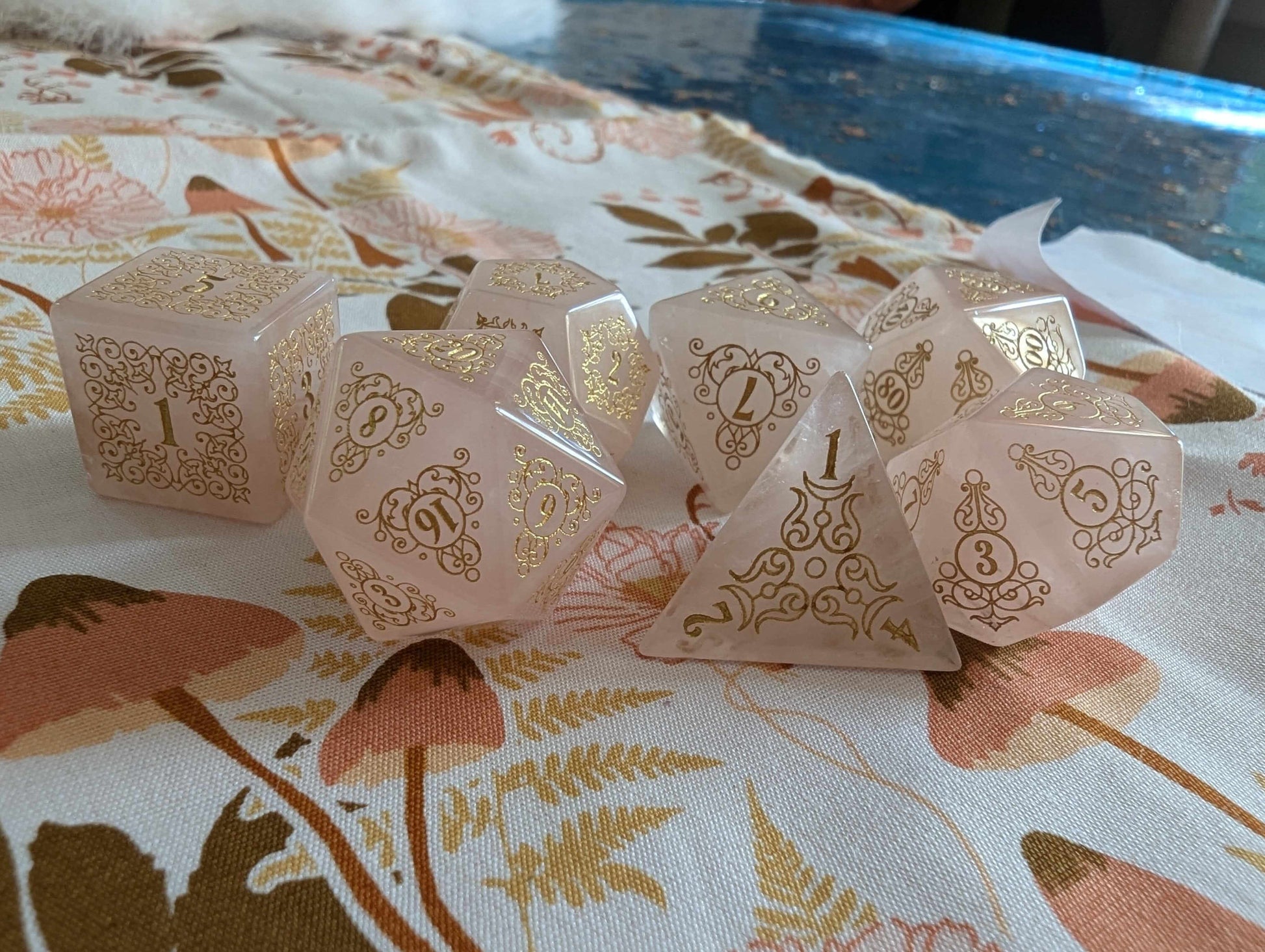 Garden Gate Rose Quartz Gemstone Dice Set with Gold Font and Intricate Designs on Floral Fabric Background