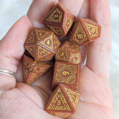 A hand holding a set of seven Geometric Goldstone dice with etched numbers, ideal for tabletop RPGs.