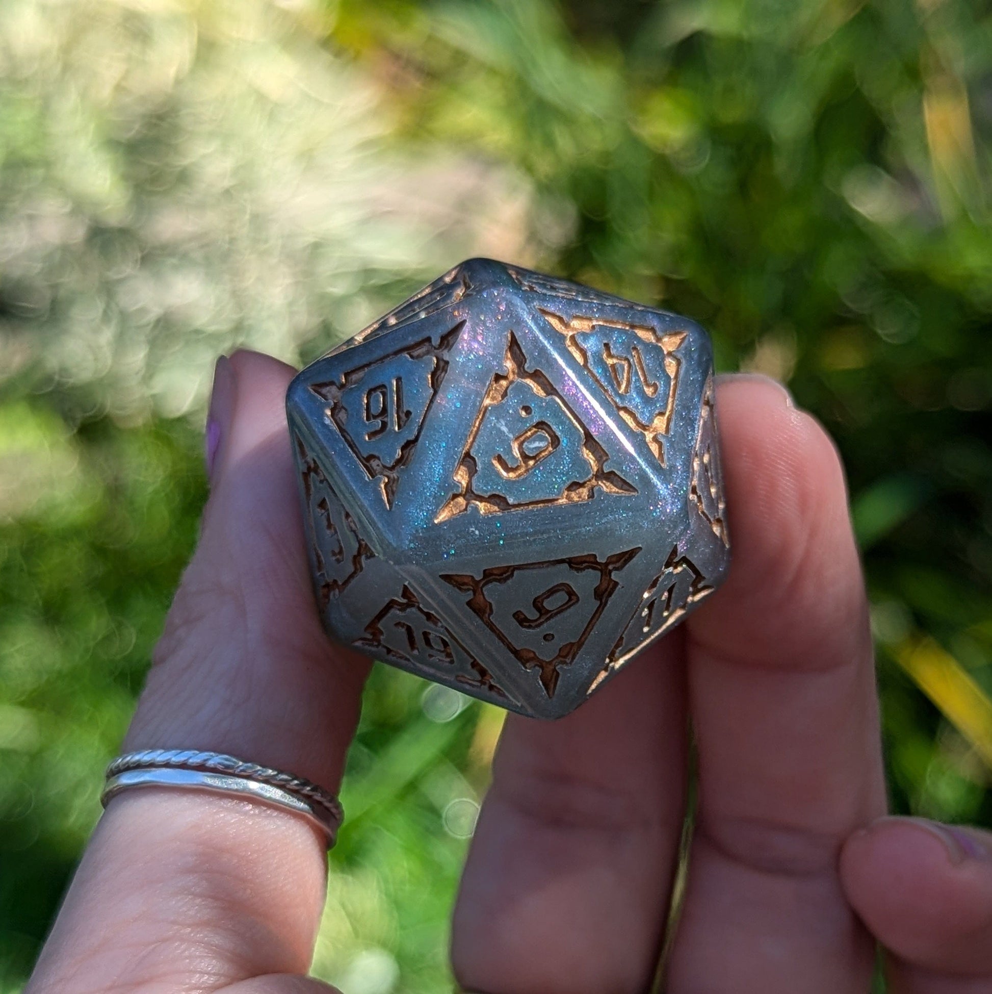 Hand holding a giant castle-themed D20 die with intricate resin details and numbers, set against a blurred natural background.