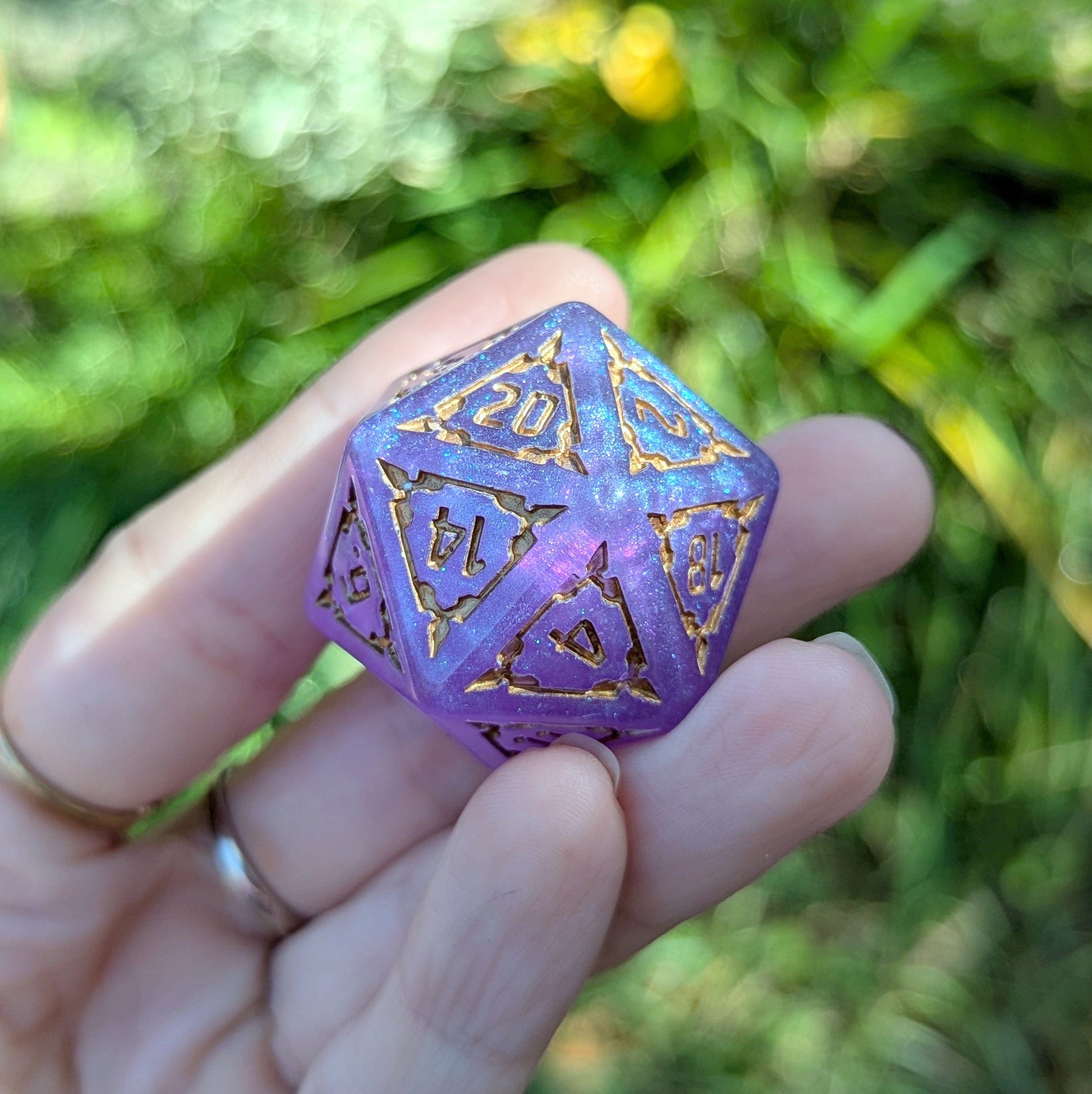 Hand holding a purple D20 die from a Giant Castle Dice set with detailed gold numbers, outdoors with greenery in the background.