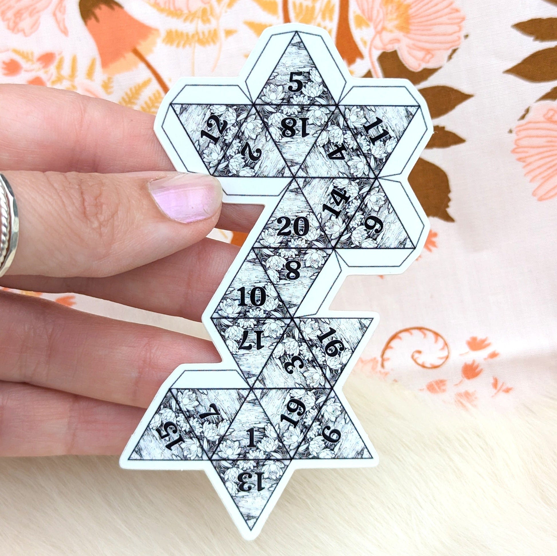 Hand holding a black and white geometric D20 sticker against a floral background, showcasing intricate patterns and numbers.