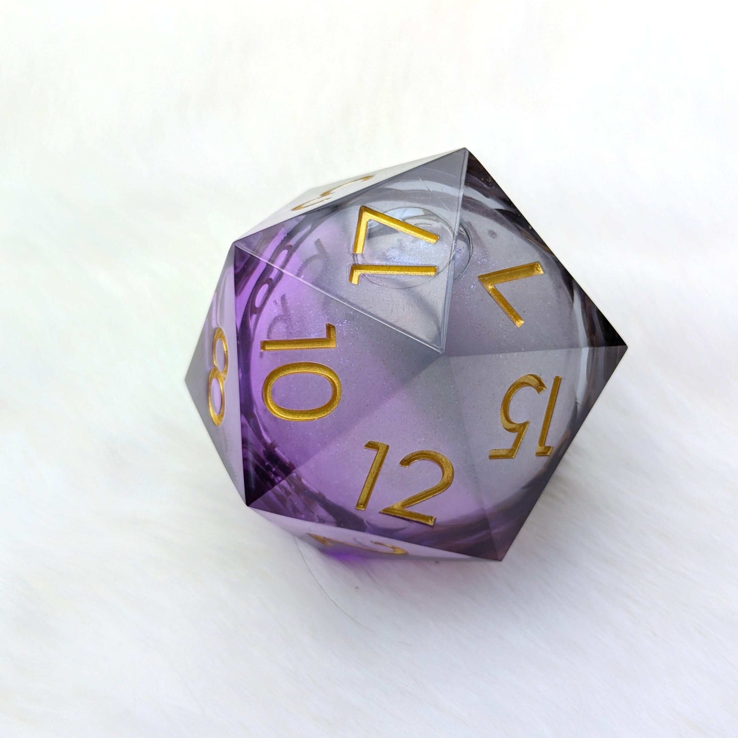 Giant D20 with purple tint and gold numbers, featuring a swirling glitter core, resting on a soft white background.