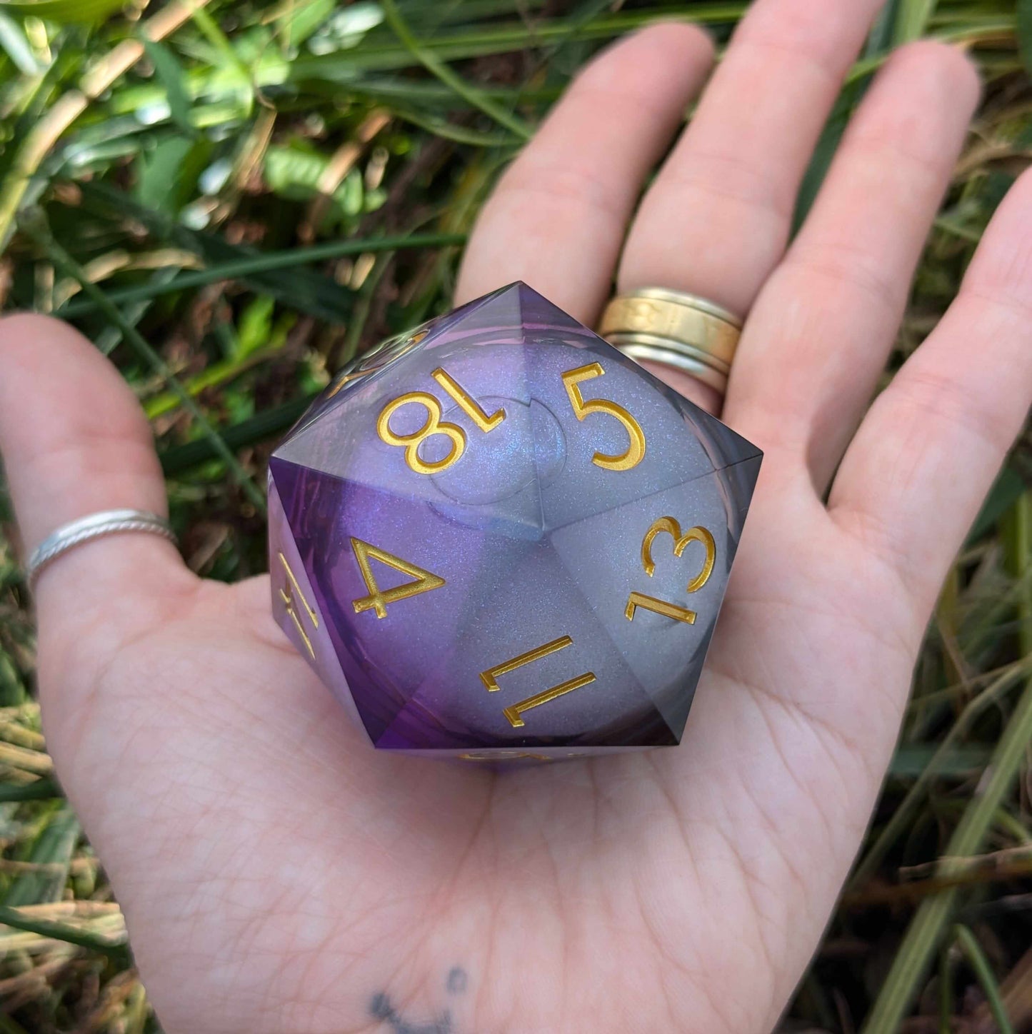 Giant D20 die in black and purple resin with gold numbers, held in hand against grass background.