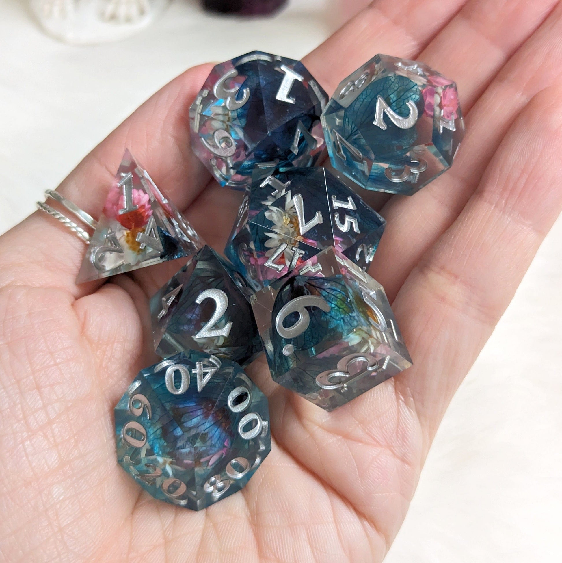 Hand holding 7 piece pink, white, and blue flower DnD dice set with silver font and embedded dried flowers.