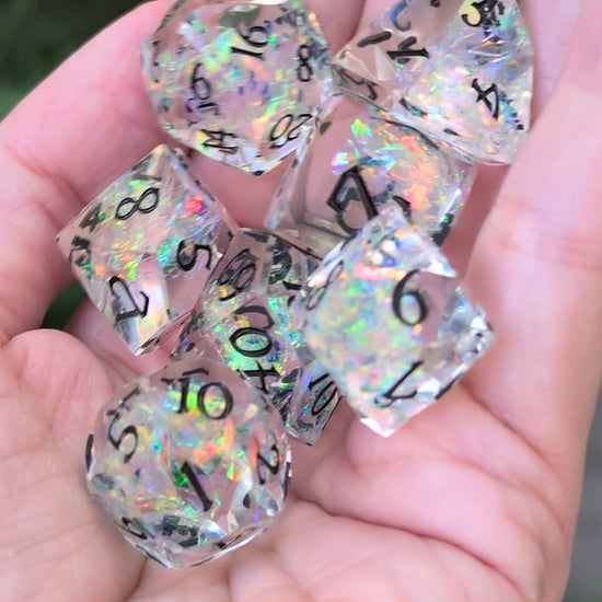 A set of clear acrylic polyhedral dice with black printed numbers and iridescent glitter being held in a hand in a video being moved around to show off the angles of the dice