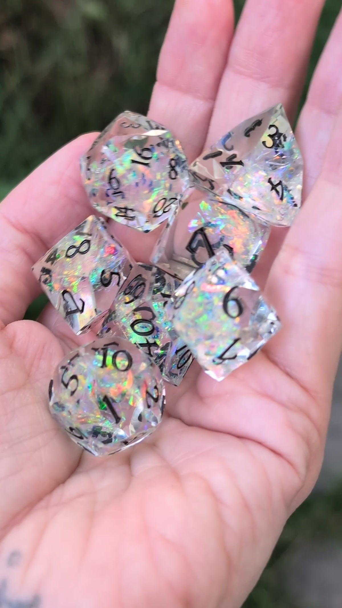 A set of clear acrylic polyhedral dice with black printed numbers and iridescent glitter being held in a hand in a video being moved around to show off the angles of the dice