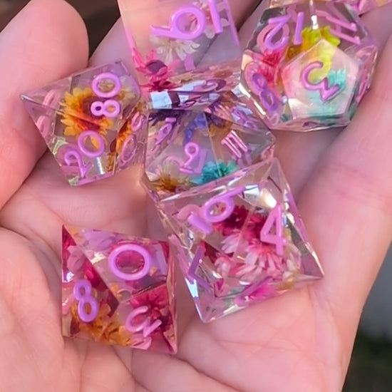 A set of transparent polyhedral dice with embedded dried flowers and purple printed numbers held in a hand to show the different dice angles in a video