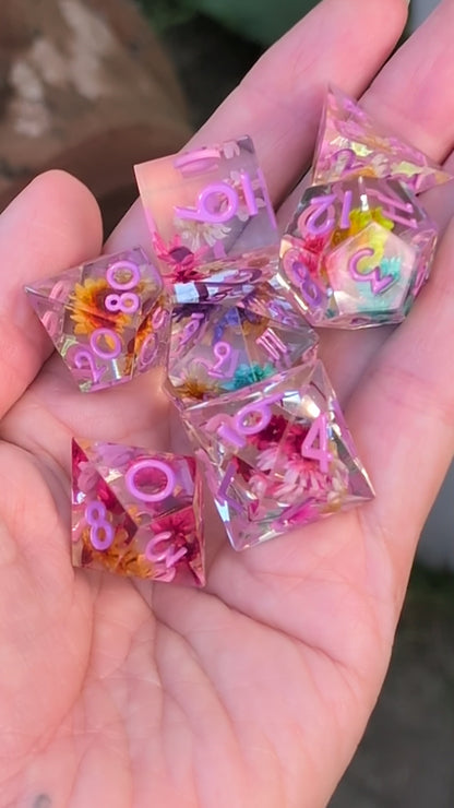 A set of transparent polyhedral dice with embedded dried flowers and purple printed numbers held in a hand to show the different dice angles in a video