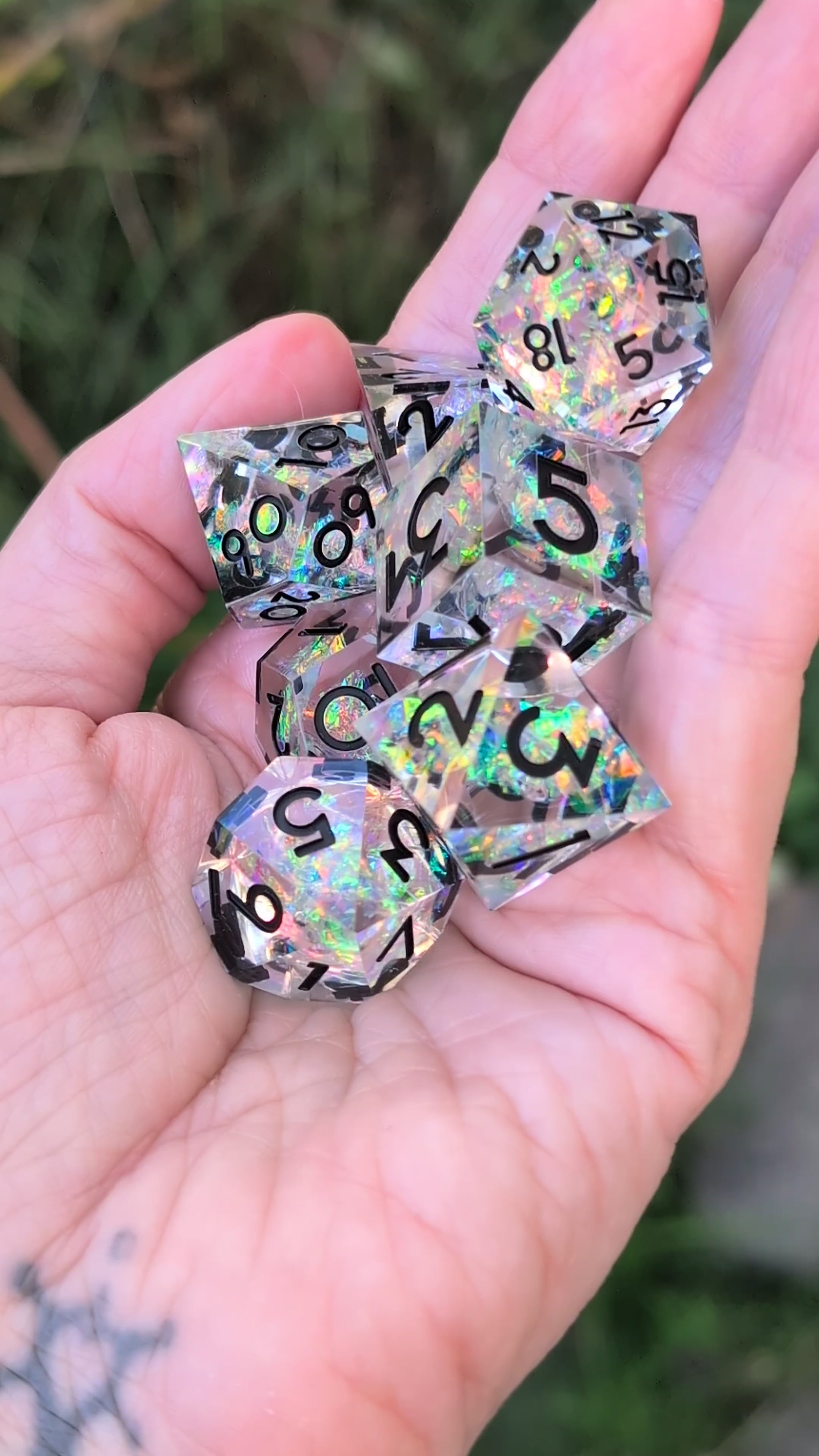 A set of holographic clear polyhedral dice with black printed numbers being show held in a hand in a video being moved around to show off the different angles of the dice