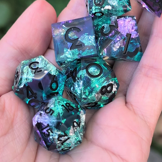 A set of translucent polyhedral gaming dice with a glittery teal, blue, and purple design being held in a hand in a video being moved around to show the different angles of the dice
