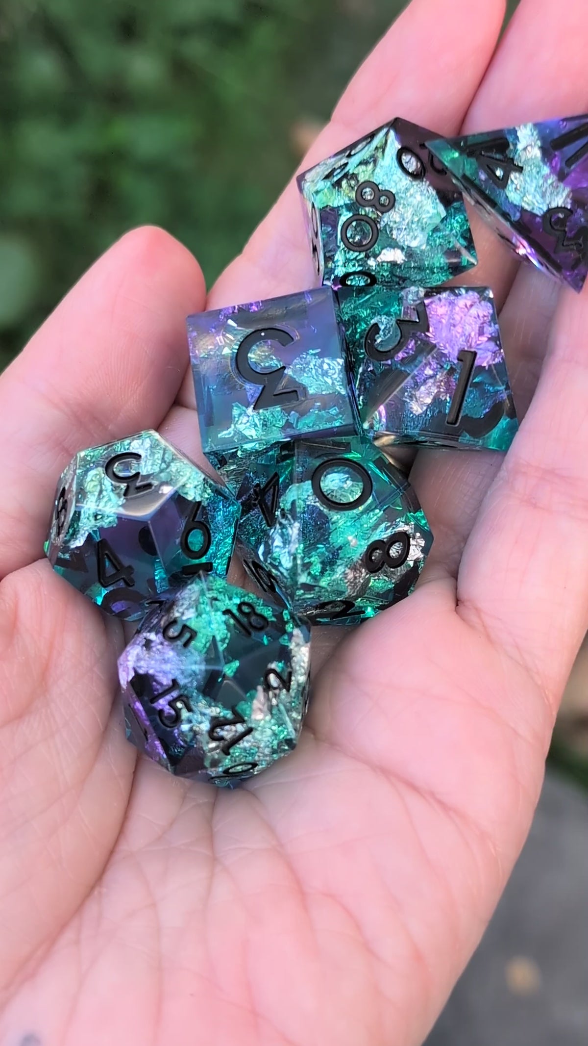 A set of translucent polyhedral gaming dice with a glittery teal, blue, and purple design being held in a hand in a video being moved around to show the different angles of the dice