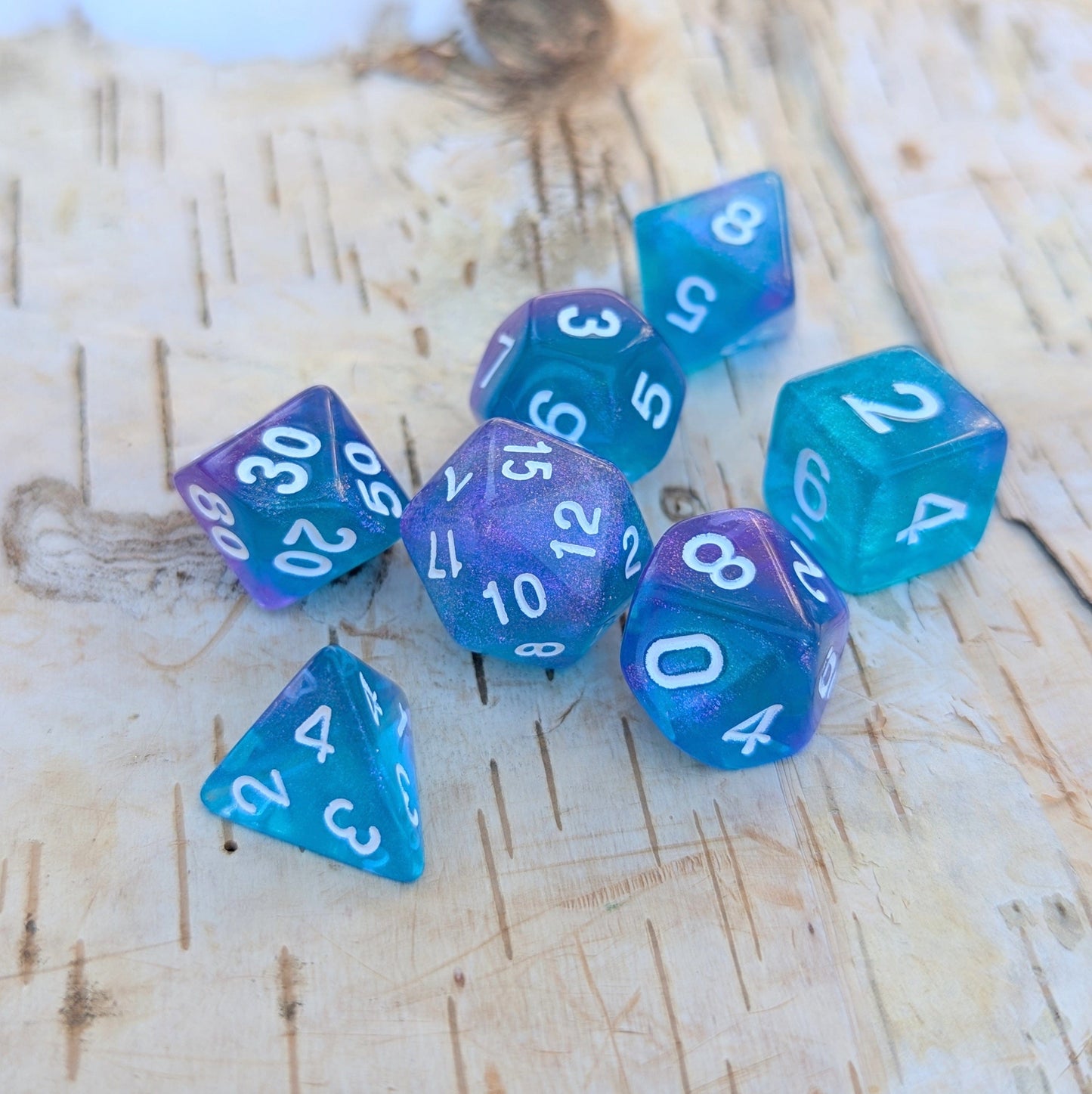 Purple and blue Will O Wisp 7-piece polyhedral dice set with shimmering glitter for tabletop role-playing games on a wooden surface.