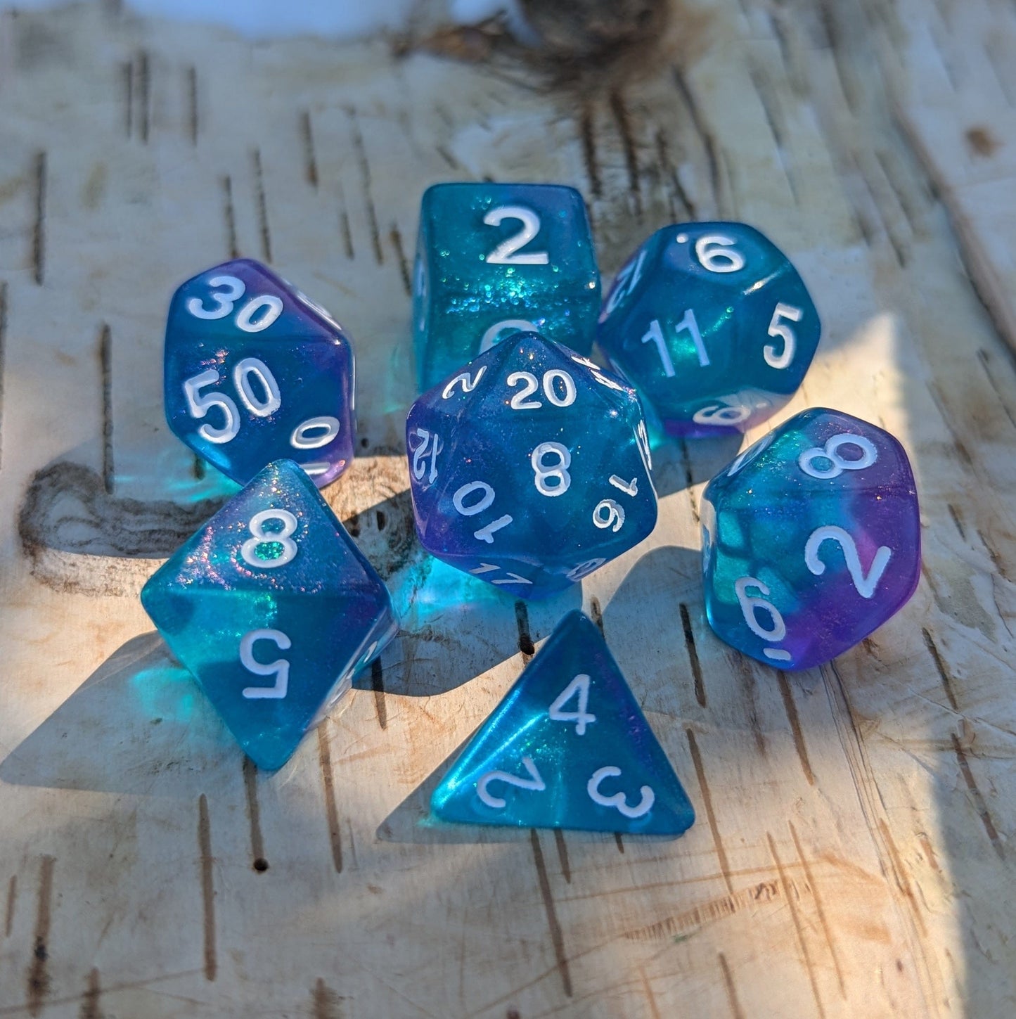 Purple and blue shimmering glitter polyhedral dice set for tabletop RPGs on a wooden surface, includes D20, D12, D10, D%, D8, D6, D4.