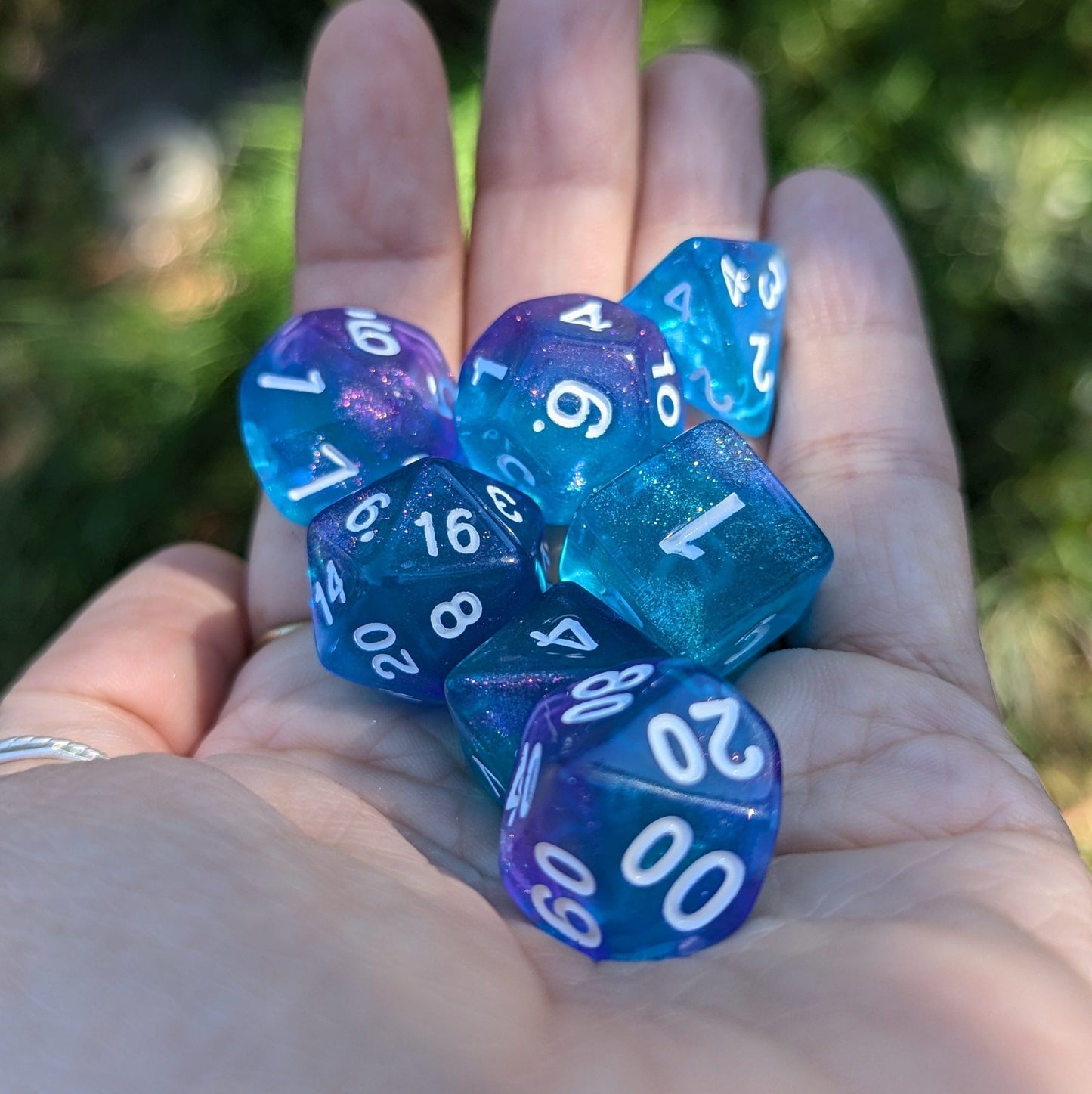 Hand holding purple and blue shimmering polyhedral dice set for tabletop RPGs like Dungeons and Dragons, includes D20, D12, D10, D8, D6, D4.